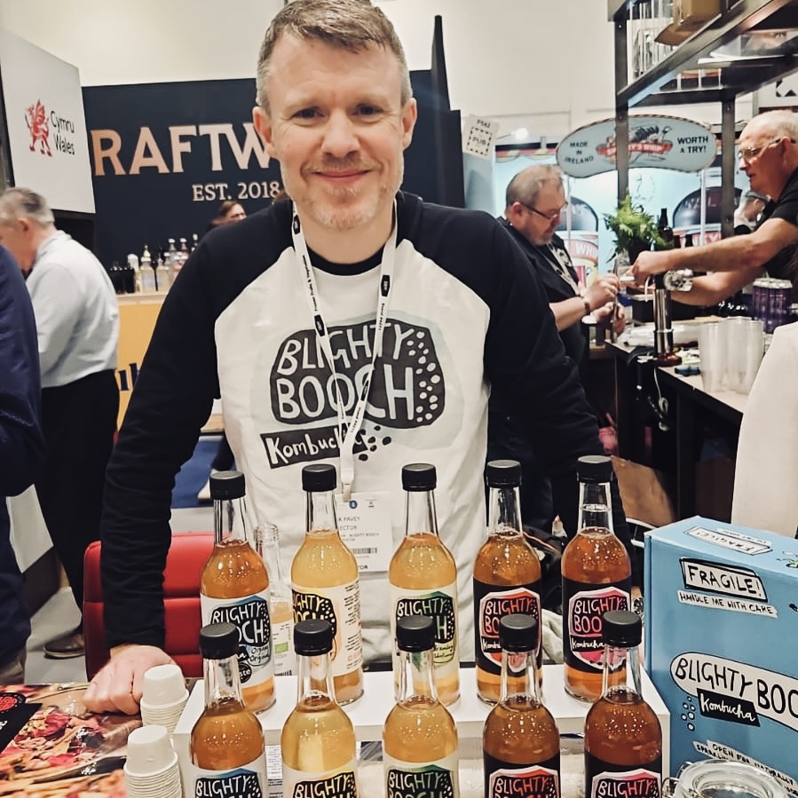Mark Pavey of Conwy Kombucha Ltd standing at a stall displaying bottles of Blighty Booch kombucha drink.